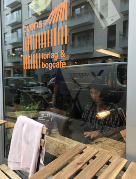Kvinde som sidder og arbejder set gennem vinduet på Byens Forlags bogcafe i Aarhus.