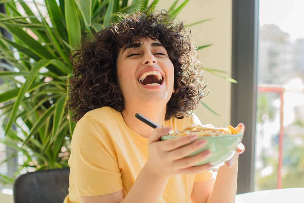 Femme souriante mangeant un bol de céréales dans une ambiance lumineuse et naturelle, symbolisant le plaisir de manger en pleine conscience et l'importance de savourer chaque repas.