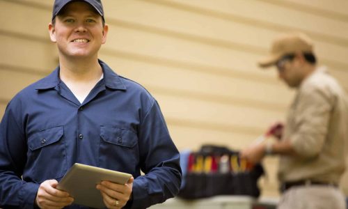 Caucasian and Latin descent repairmen work on a home's air conditioner unit outdoors. One foreground is using a digital tablet. Other man in backgroud is checking unit using the tools in his tool bag.  They both wear uniforms.  Repairmen, electricians, inspectors, service workers.