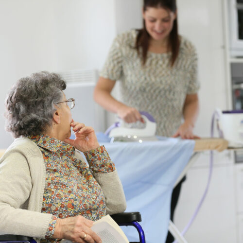 Elderly woman reading book while home helper irons laundry