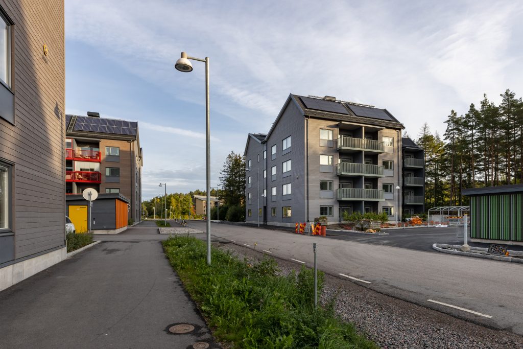 Flerfamiljshus med fyra våningar samt grå och bruna träfasader på varsin sida av en bilväg med gång-/cykelbana.