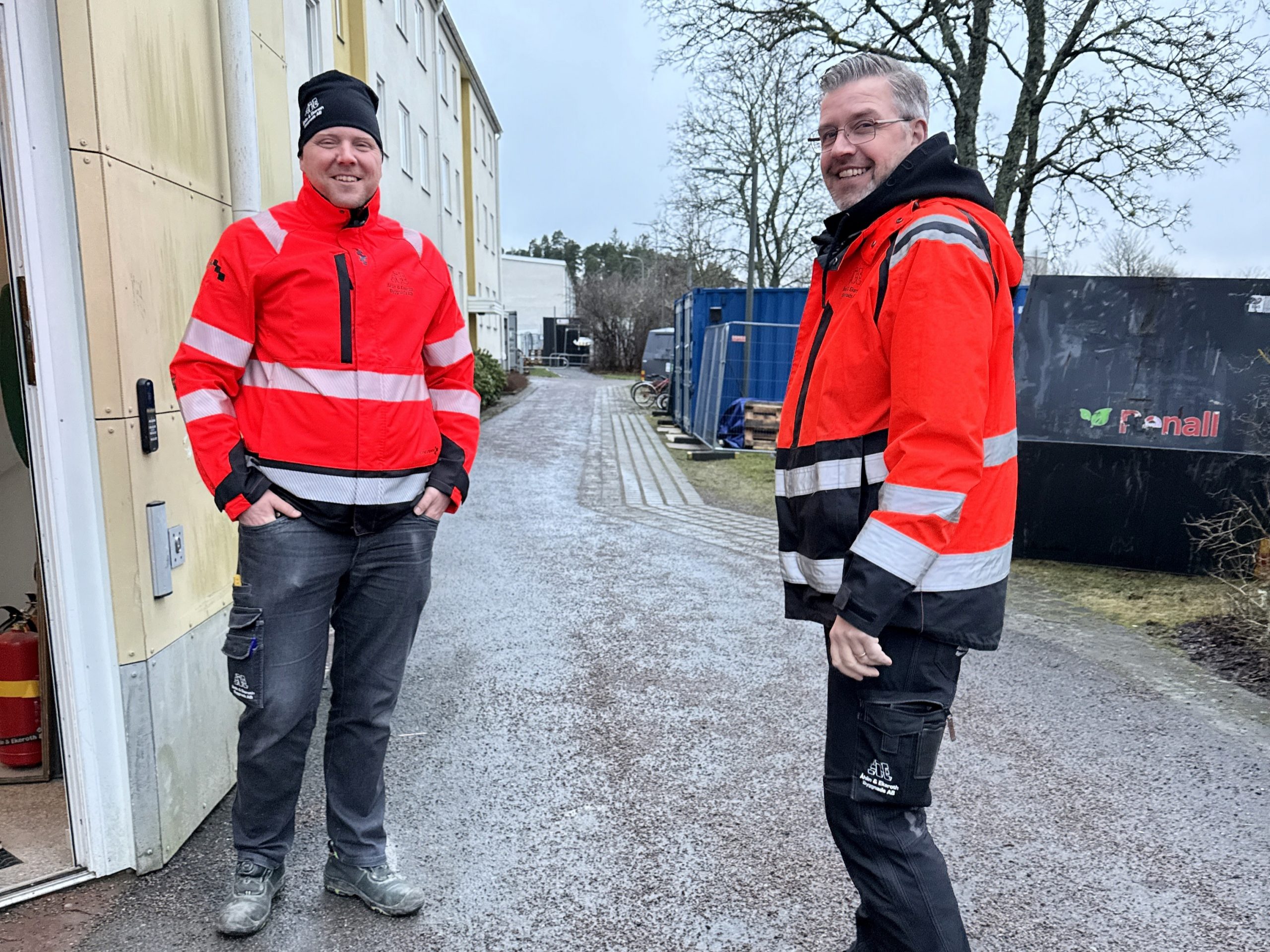 Produktionschef Anders Ringdahl och produktionsledare Anders Falkestål från Åhlin & Ekeroth Byggnads AB utanför ett bostadshus på Kullborg i Söderköping. De har orangea varseljackor på sig och ser glada ut.