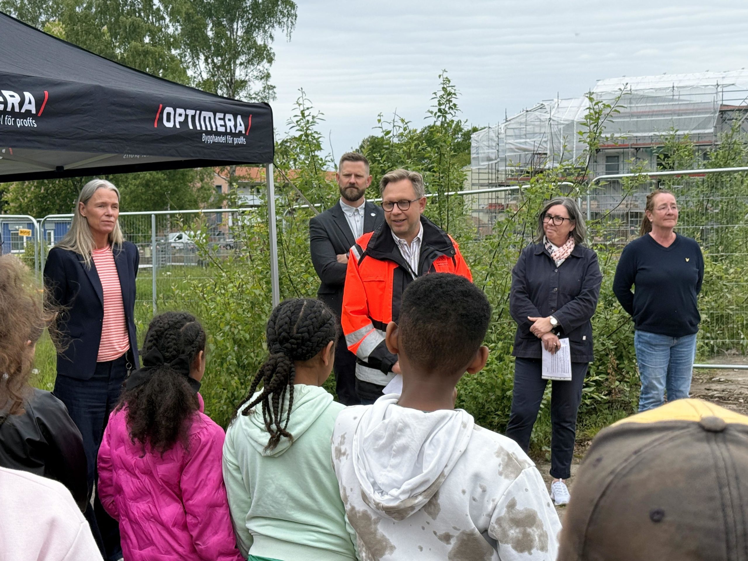 Åhlin & Ekeroth Byggnads AB:s affärsområdeschef Magnus Welin Harrström håller tal inför en skara skolbarn vid första spadtaget för Nya Vätternskolan i Motala. Vi ser Magnus vänd mot kameran iklädd orange varseljacka. Barnen står med ryggarna mot kameran och tittar på Magnus. Fyra andra vuxna står utspridda runt Magnus.