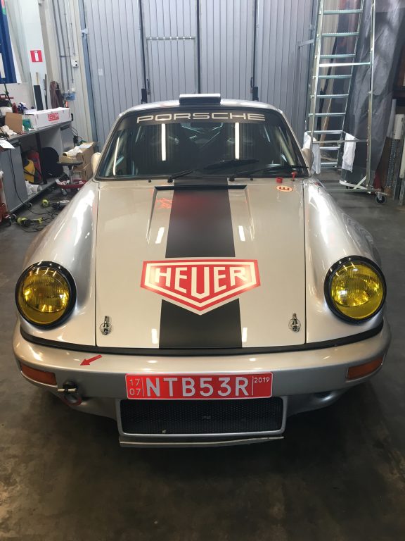 En silvrig sportbil framifrån med texten Porsche längst upp på framrutan. På huven syns en bred svart rand och däröver en vit text med röd bakgrund: "Heuer".