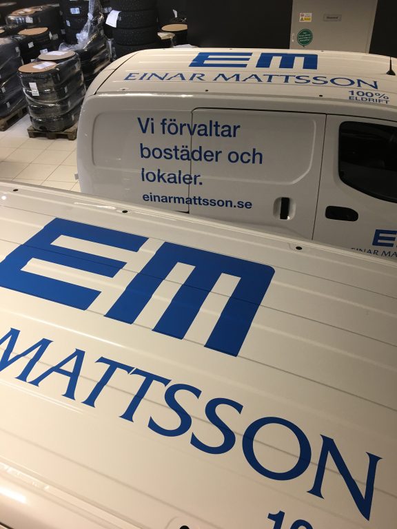 Två vita transportbilar, fotograferade något uppifrån. Har blå texter på taket där det står EM Mattsson, och på ena bilens sida står texten "Vi förvaltar bostäder och lokaler. Einarmattsson.se".