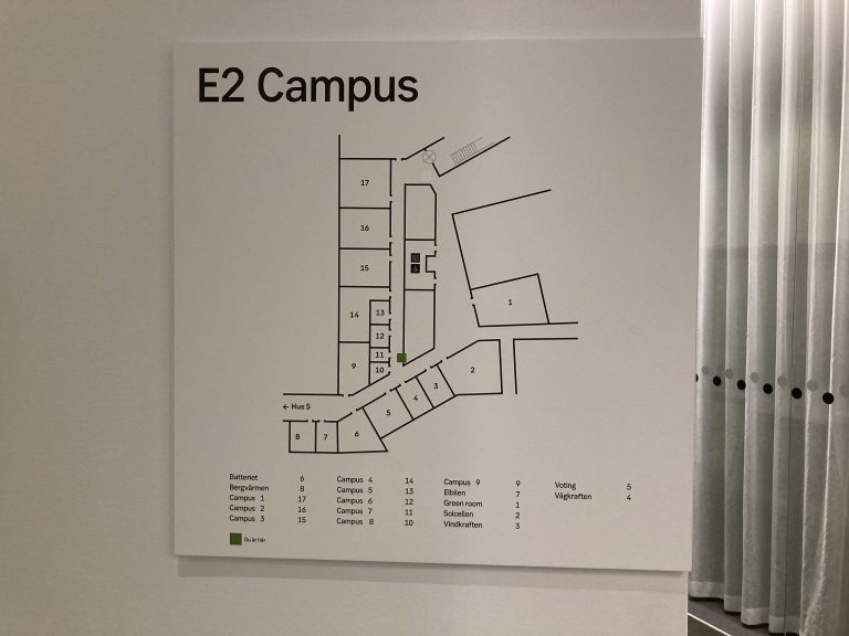 En vit vägg med en vit skylt, på vilken en karta över ett campus syns. Texten E2 Campus står i vänster överkant i svart och kartan är uppmärkt med siffror för de olika lokalerna och förteckning i nederkant.