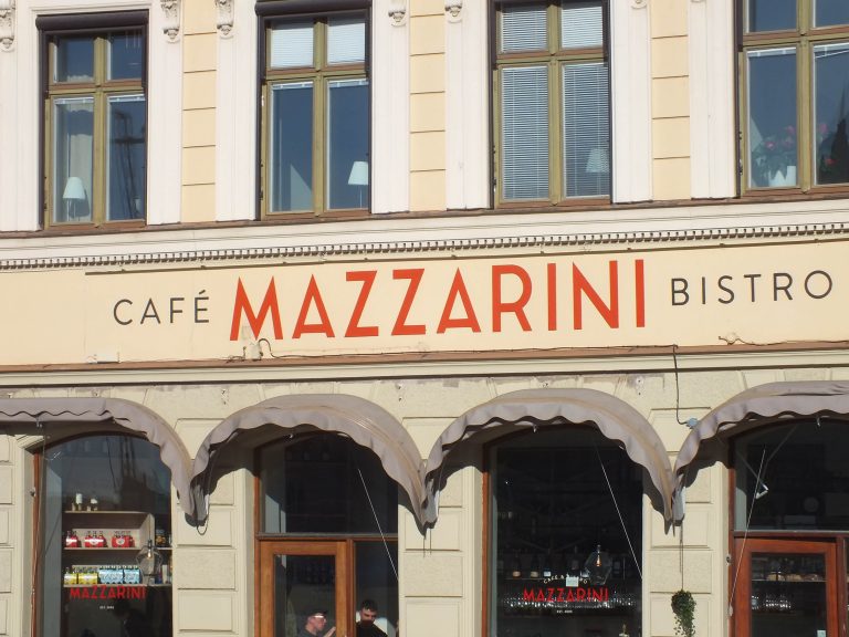 Husfasad med butik, över vilken en ljusgul yta har den målade texten "Café Mazzarini Bistro". Ordet MAZZARINI är i rött och övrigt i mörkgrå.