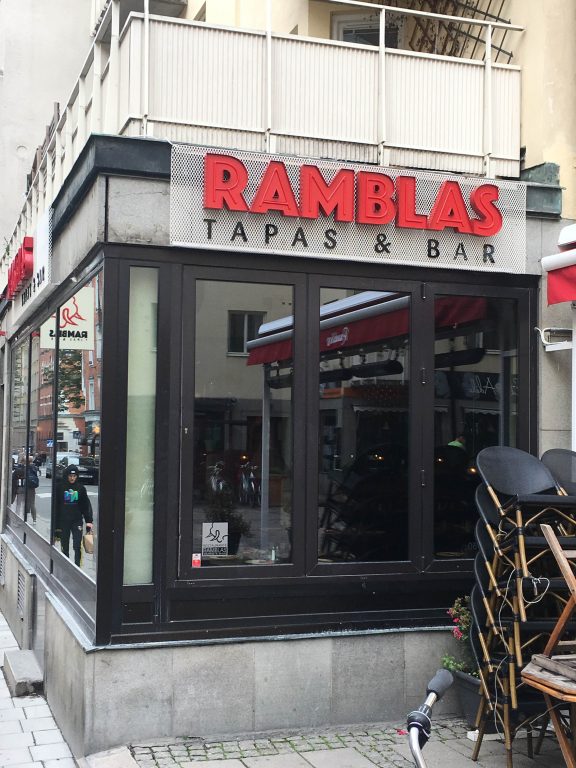 Butiksfönster med väggskylt ovanför. Skylten är en neonskylt med rödlackade metallformar för bokstäverna som utläses "Ramblas", och därunder i svarta, mindre, bokstäver "Tapas & bar".