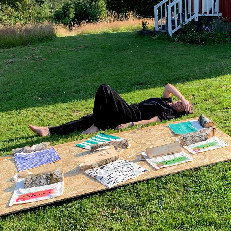Konstnären Alinde Jormfeldt ligger i gräset och vaktar handtryckta ekologiska textil väskor som torkar i solen