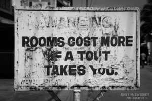 Old black and white street sign in Mamalapuram India warning "Rooms Cost More if A Tout Takes You"