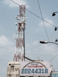 Street sign in India saying "Contact Rainbow" with street lamps and tower.