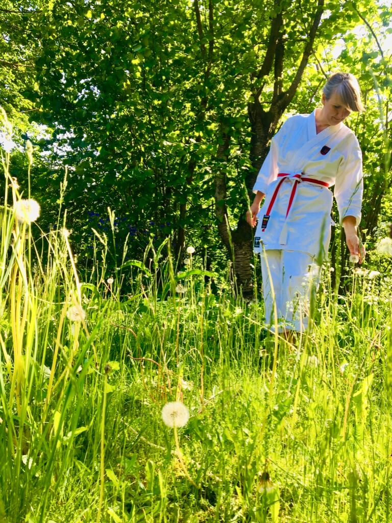 Anja Dalby i naturen, grundsten i en spirituel praksis. Hun står i højt græs med afblomstrede mælkebøtter i sin karatedragt.