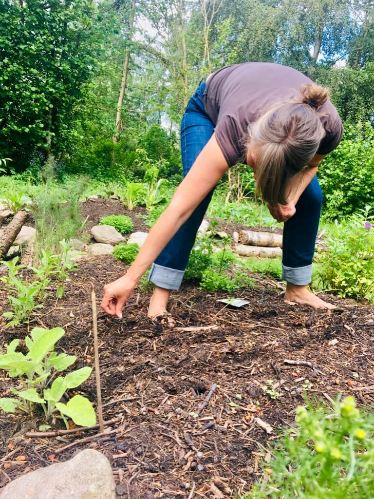 Anja Dalby: Min mor-side vs. intuitionen. Anja står i sin have med fødderne i jorden i et bed. Hun bøjer sig ned og planter frø.