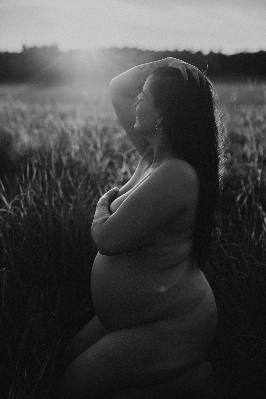 Gravid kvinna fotograferad i högt gräs, sittandes naken i solnedgången under gravidfotografering i Stockholm