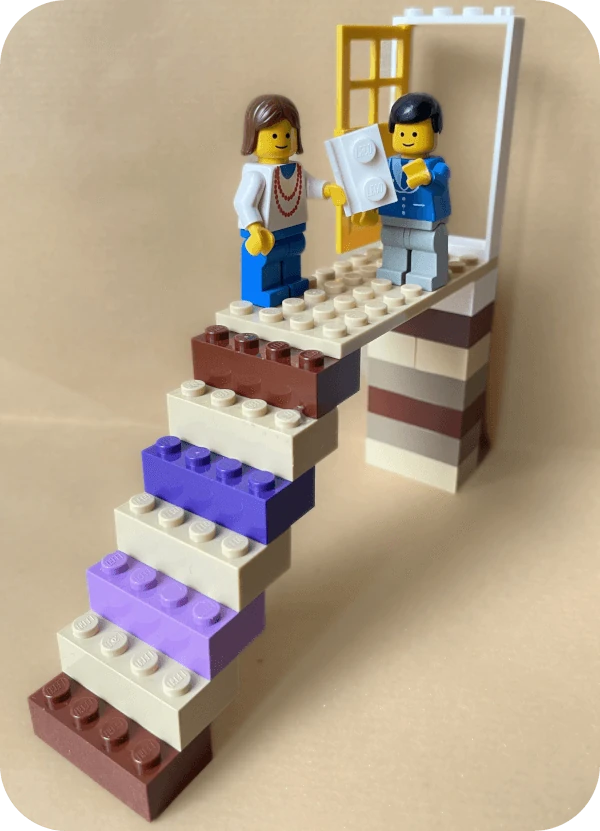 Coach für berufliche Neuorientierung Hamburg, Lego-Szene: Auf einer Plattform am Ende einer Treppe überreicht ein Mann einer Frau einen Arbeitsvertrag, beide stehen vor einer weit geöffneten Tür