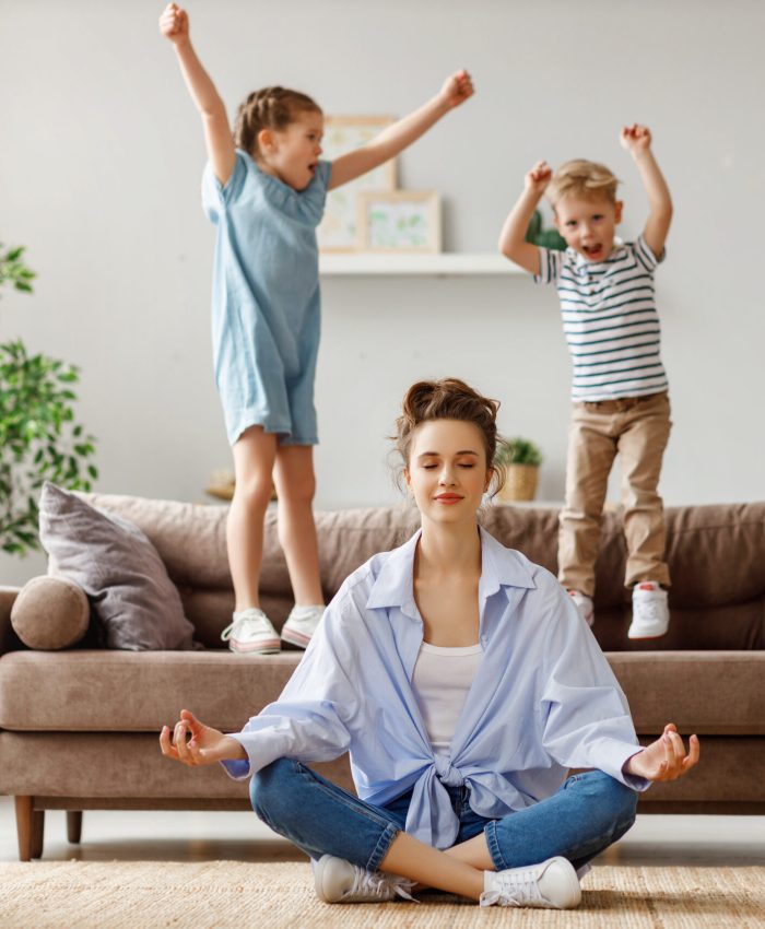 Happy mother with closed eyes meditating in lotus pose on floor trying to save inner harmony while excited children jumping on sofa and screaming in light spacious living room