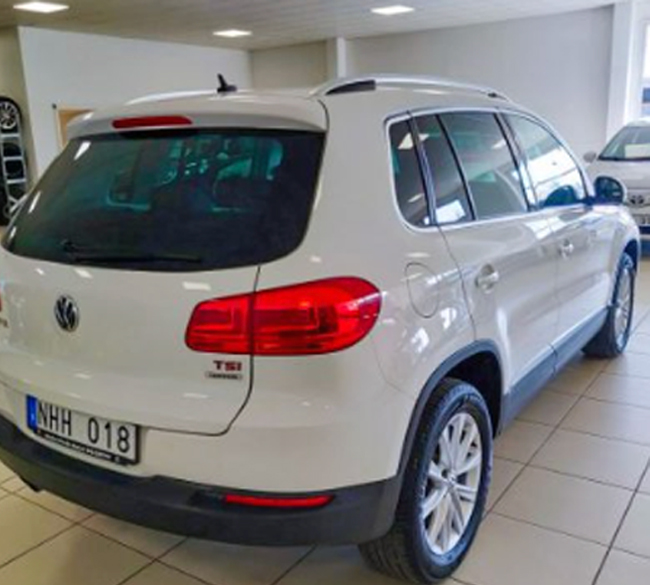 Vit Volkswagen SUV sedd bakifrån i en bilhall, parkerad på klinkergolv med registreringsskylt "NHH 018" synlig och flera andra bilar i bakgrunden.