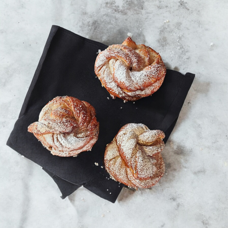 På bilden ligger det 3 stycken kardemummaknutar som är pudrade med florsocker. Dom ligger på en svart kökshanduk och representerar våran produkt kardemummaknut.