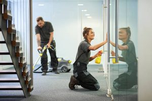 a female cleaning contractor is polishing the glass partition offices whilst In the background a male colleague steam cleans an office carpet in a empty office in between tenants.  .The female is smiling .