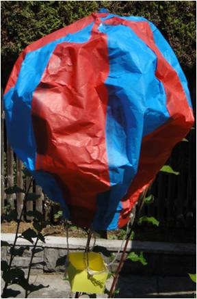 Selbstgebauter Heißluftballon in blau-rot. Er hängt in der Luft mit einem gelben Korb.