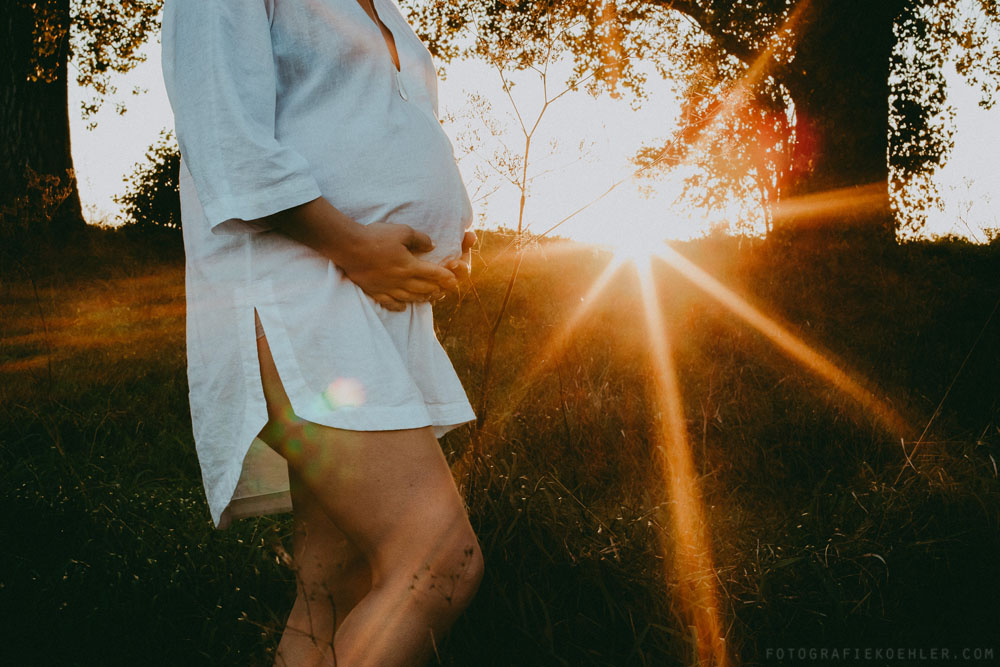 eine schwangere Frau steht draussen in der Natur vor einem kleinen Hügel, wo im Hintergrund die Sonne langsam hinterm Horizont verschwindet.