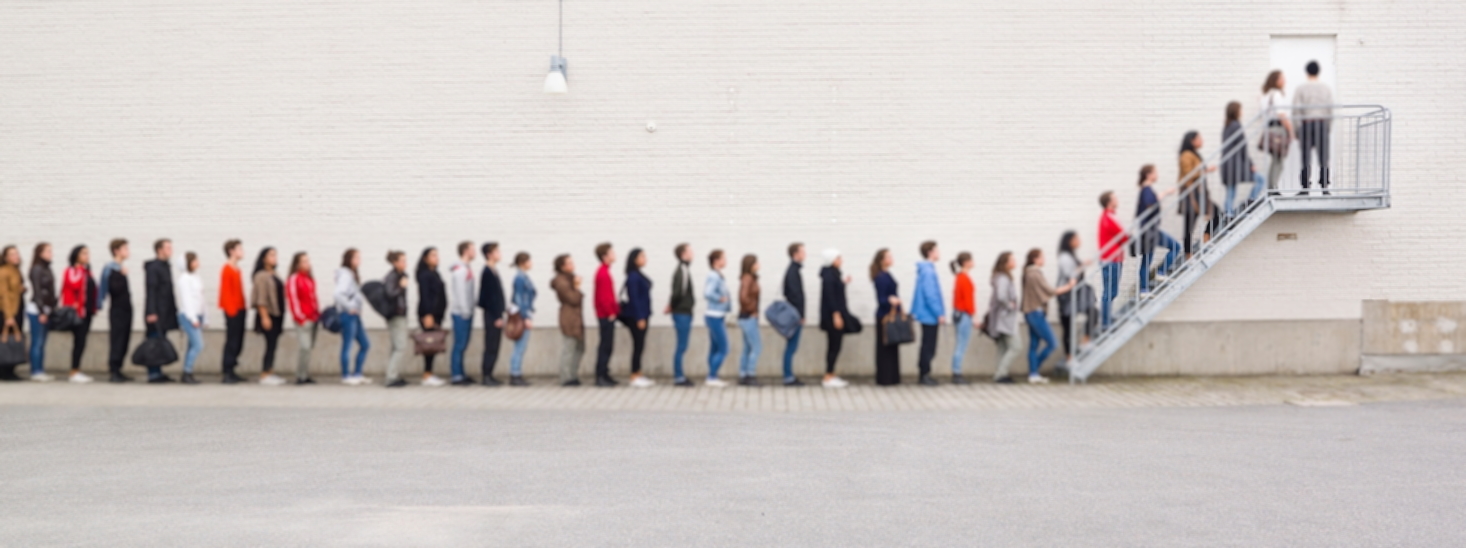 People are standing in a line along a white wall to reach the door on top of the stairs on the right.