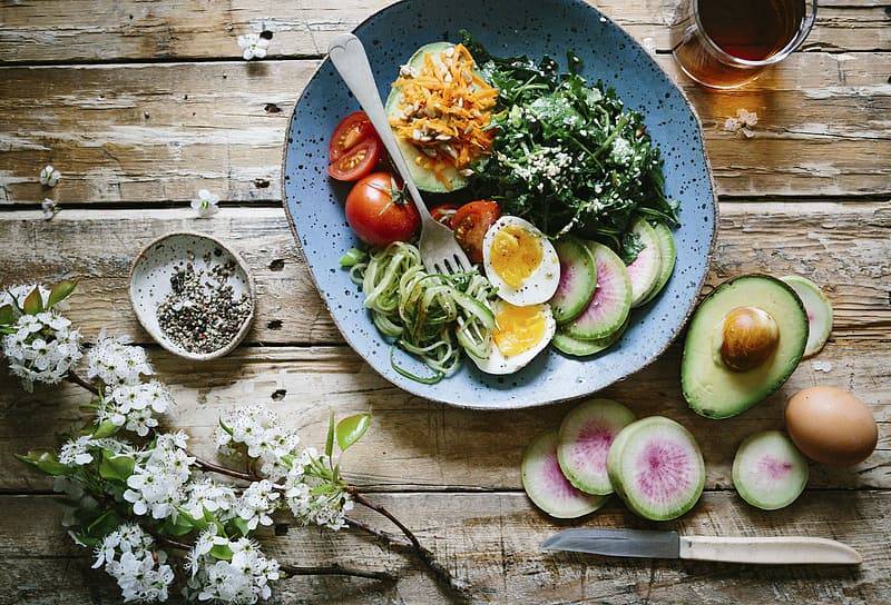 Blå keramiktallrik med grönsaker och ägg, avokado ligger på sidan av på ett gamalt rustik träbord med blommor och ett glas dryck. 
Balansera ph-värden med hjälp av kosten.