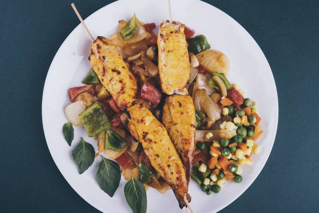 Vit tallrik på blått bord, med två kycklingspätt ovanpå grönsaker, varm sallad och grillade grönskaer. Det ligger ärtor, majs, morötter, grillad varm lök, paprika och gröna blad. Lågkolhydratskoster