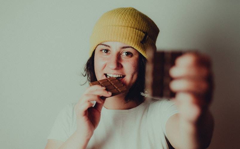 Tjej med brunt kort hår i page och gul mössa på huvudet håller choklad. En chokladkaka i ena handen som hon håller upp framför kameran. Den handen är ur fokus och suddig. Den andra handen håller en annan bit choklad som hon har i munnen, med ett leende och tittar in i kameran.