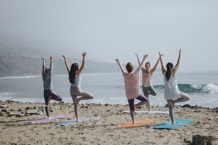 grupp på 5 personer gör yoga på en strand. De står på varsin yogamatta i "trädet" med ena benet i knävecket på det andra benet samt båda armarna rakt upp ovanför huvudet. Yogalärarens står vänd mot de deltagande. Hon står med ryggen mot vattnet och de andra står vända mot vattnet. Det är en liten våg vid strandkanten. Rolig träning