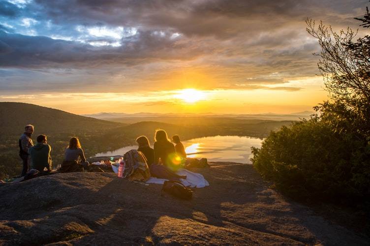 en grupp med 6 personer sitter på en bergstopp vid solnedgången och tittar ut över vyn med berg, skog och sjö. Solen ligger mellan molnen vid horisonten. Gruppen av kompisar har tagit med sig fika och mat samt filtar som de sitter på. Vid sidan är låga buskar. Naturen bidrar till hälsa