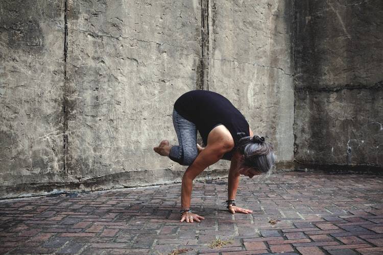 kvinna med svart linne och gråa träningsbyxor håller på med yoga och gör "crow pose". Hon balanserar på händerna på ett stengolv med stenväggar runtomkring. Håret är mörkt med gråa slingor och uppsatt i en tofs. På armlederna har hon flera armband.