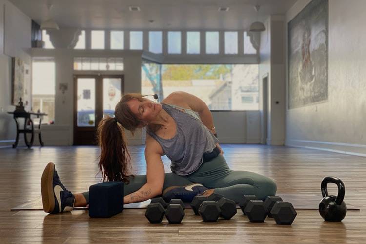 Kvinna strechar på en yogamatta med styrketräningsvikter framför sig, dumbells, lösa vikter. Hon har ett par gröna träningsbyxor på sig och ett lila linne, håret är brunt och uppsatt i en tofs. Runt huvudet har hon en mikrofon. Rummet hon sitter i är tomt, ett stort rum med fönster vettande ut mot en gata. Det är ingen annan i bilden.