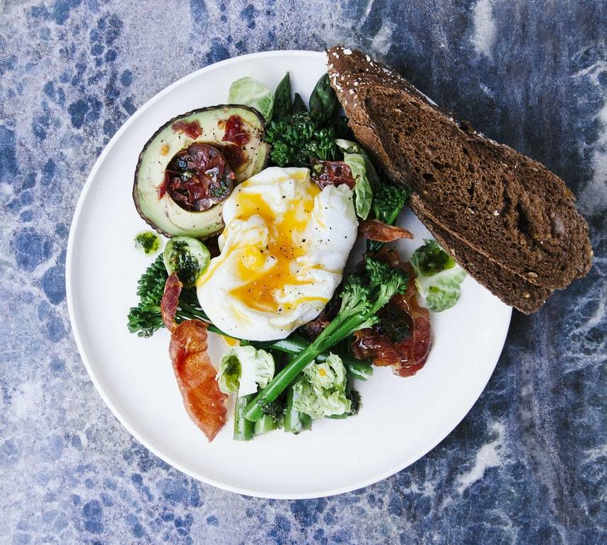 Vit tallrik med grönsaker, sallad, halv avokado, ägg och mörkt bröd. Tallriken står på ett blått bord. Ägget ligger ovanpå grönsakerna och brödet skuret och ligger på sidan av tallriken.