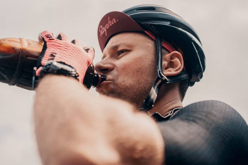 Man dricker ur en plastflaska när han cyklar under träning eller tävling. Bilden är tagen på den övre delen av mannen med huvud och delar av överkroppen. På huvudet har mannen en keps och ovanpå en hjälm. På händerna han röda cykelhandskar.