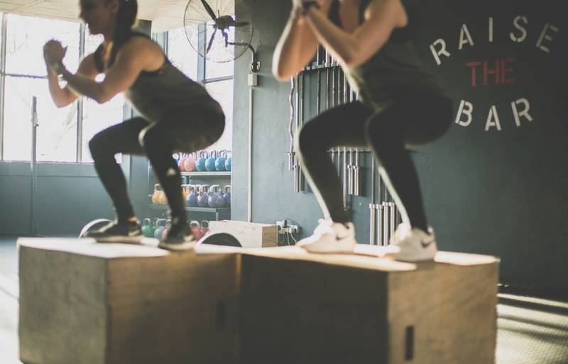 Två kvinnor tränar styrketräning med trälåda. De gör benövningar på lådan, står i squat med händerna ihop framför sig. Tjejerna har svarta träningskläder på sig, tights och linne. Ena tjejen har vita träningsskor och den andra svarta träningsskor. I bakgrunden ser man kettlebells i olika vikter som står uppradade på hyllor. Väggen bakom tjejerna är svart med texten "raise the bar". Det hänger en fläkt på väggen bredvid samt stänger utan vikter hänger uppradade på väggen.