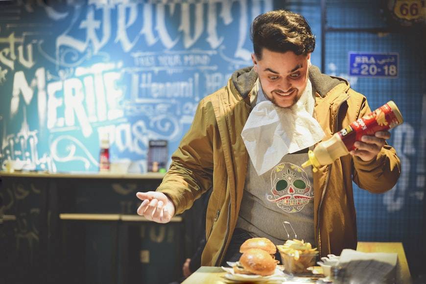 Glad kille med servett under hakan och ketchupflaska i handen. Han tittar glatt ner på sin tallrik med hamburgare och pommes frittes. Killen befinner sig på en hamburgerrestaurang och har precis blivit serverad maten. Han står vid bordet, ensam. Hormoner styr hunger och sug