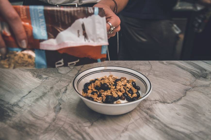 Kvinna häller granola i en skål med yoghurt och blåbär. I skålen ligger det redan granola och kvinnan håller påsen ovanför. Skålen står på en köksbänk. Snacks vid hunger