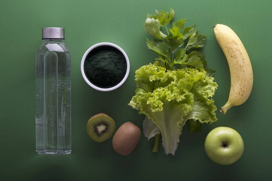 En vit liten skål med spirulina står på ett grönt bord med frukter, grönsaker och en flaska. Det ligger en banan, salladsblad, persilja, kiwi och grönt äpple på bordet. örter som kan öka träningsprestation