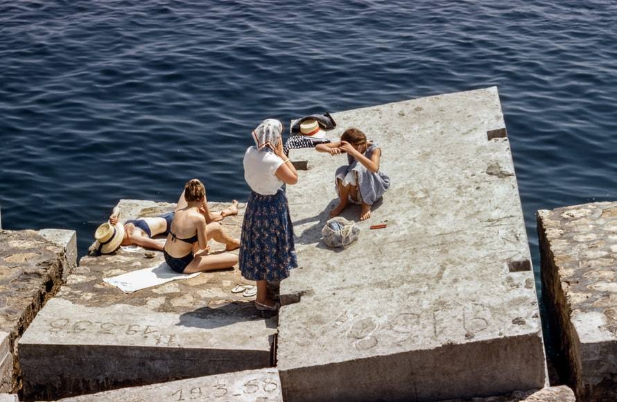 Solning på klippblock. Tre kvinnor och en flicka solar på stora klippblock av betong. Två kvinnor ligger ner med bikini och en med hatt över huvudet. Flickan sitter med klänning på och fixar med sitt hår. Den tredje kvinnan står upp med kläder på och en sjal över huvudet. D-vitamin av solen.
