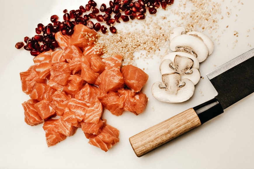 Skurna bitar av röd lax och skivade champinjoner ligger på ett vitt underlag. Bredvid ligger en kniv, kärnor av granatäpple samt små frön, möjligen quinoa.Livsmedel med D-vitamin