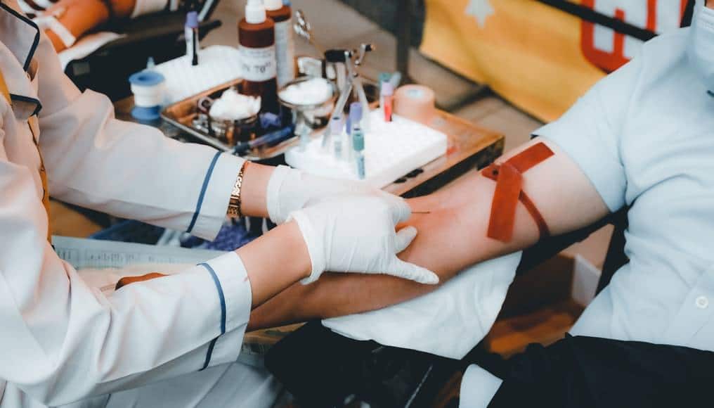 Blodprov tas av en sjuksköterska med vita plasthandskar. Det sitter en kvinna med mask och kortärmad vit t-shirt och en röd rem runt armen och med en spruta i armvecket. I bakgrunden syns diverse sjukmaterial på ett litet bord.