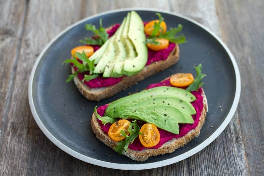 Skivor av smörgåsar med röda rödbetsröra, skivad avocado, ruccola och coctailtomater i skivor. Smörgåsarna ligger på en grå tallrik som står på ett mörkt träbord. Hälsosam kost med grönsaker för att förebygga cancer