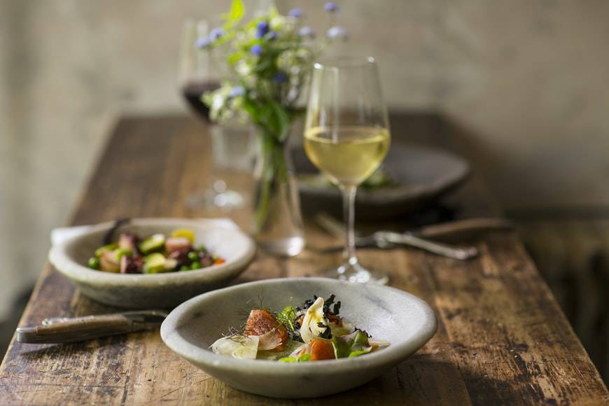 Tre gråa djupa tallrikar med mat står på ett träbord med två glas vin. Ett glas rött vin och ett glas vitt vin. Mellan glasen står en liten blombukett i en glasvas. Besticken ligger bredvid tallrikarna.