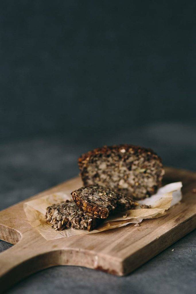 Fullkornsbröd med mycket nötter och frön. Brödet ligger på en skärbräda av trä och ett par skivor är uppskurna och ligger på en bit bakplåtspapper framför limpan. Brödet är mörkt och med mycket fibrer och fullkorn