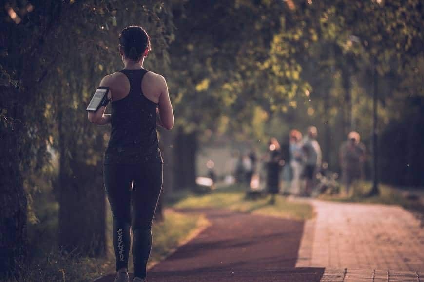 Kvinna ute på joggingtur i stadspark. Hon har på sig ett svart linne och svarta långa byxor. Runt armen har hon en ipodhållare och hon har håret uppsatt i en hästsvans. Hon springer på en asfalterad väg med kullersten på sidan och träd. I bakgrunden syns en grupp människor. Bilden är tagen bakifrån av kvinnan. Förebygga cancer med fysisk aktivitet