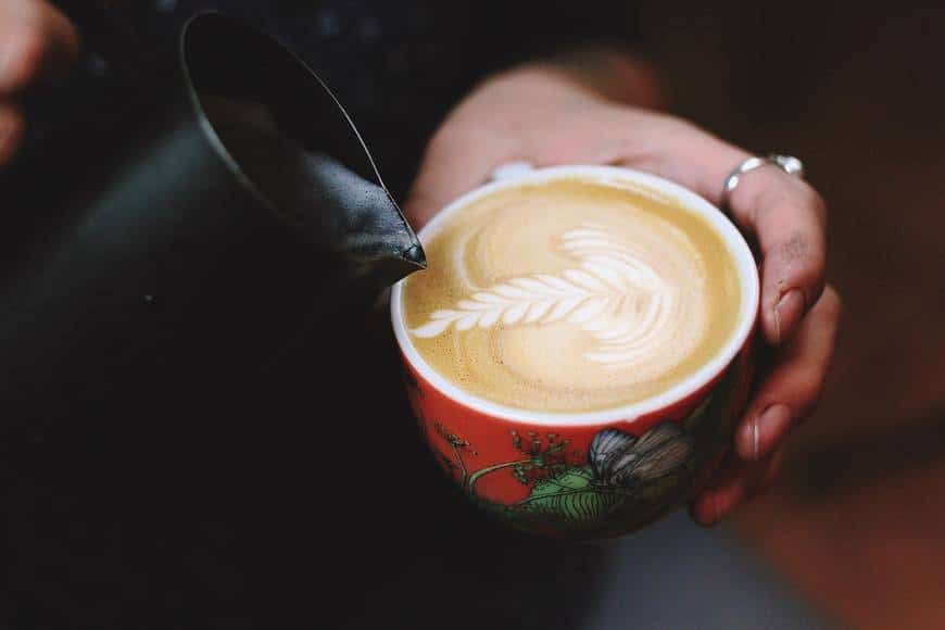 Kvinnlig barista häller upp mjölk i capuccino. Hon häller den så att mjölken bildar ett mönster av en blomma. Koppen är röd och mönstrad med blommor. Kvinnan har ringar på sina händer. Kortet är taget så att man enbart ser händerna, koppen och mjölkkannnan. Glad av kaffe och koffein