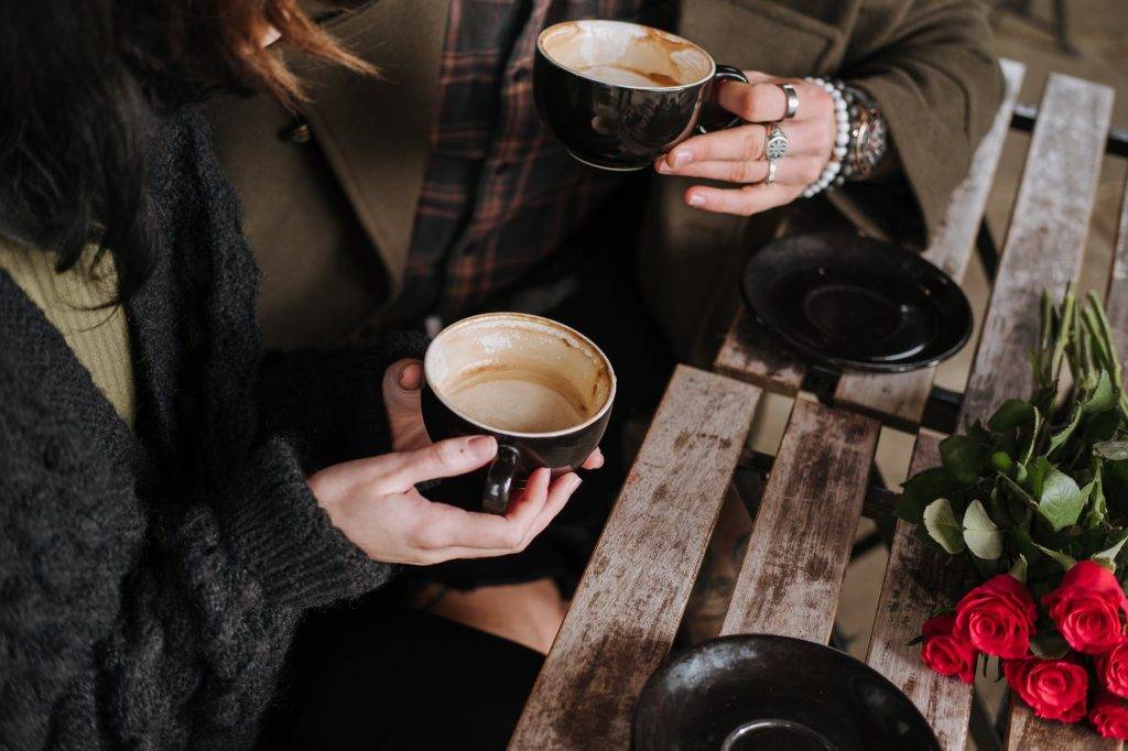 Två kvinnor sitter och dricker kaffe tillsammans på vad som ser ut som ett kaffe med träbord för utomhusbruk. På bordet ligger en bukett röda rosor och det står två svarat kaffefat. Kvinnorna håller en varsin svart kaffekopp i handen som är uppdruckna. Bilden är tagen så att man inte ser ansiktena på kvinnoran utan framförallt händerna och kaffekopparna. Ena kvinnan har många smycken runt sin handled och ringar på fingrarna. Glada av kaffe