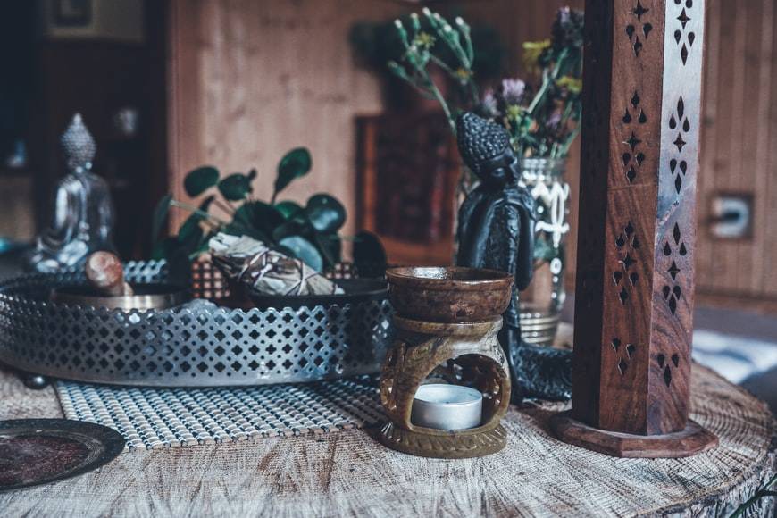 Aromalampa i brun keramik med värmeljus står på ett träbord med andra saker. Bordet är som ett altare med statyer, buddhastatyer, rökelse och rökelsehållare, kristaller, salvia i bunt, en vas med blommor och sångskål. Det står en silverbricka med saker i sig.