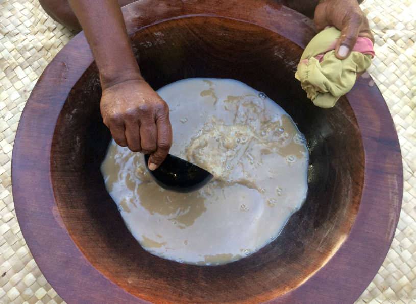 Kvinna gör te på traditionellt vis av roten kava kava, piper methysticum. Bilden är tagen ovanifrån där man ser ett stort fat i trä med te av kava kava samt kvinnan som håller en skopa i teet. I andra handen håller hon en tygbit. Upplyftande mot nedstämdhet under PMS